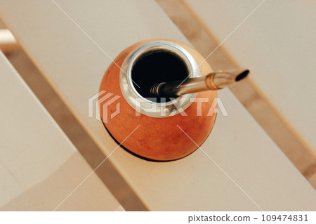 Mate calabash with bombilla on a light warm background. Healthy infused drink, classic Yerba mate tea on a table top view. Hot beverage on cafe terrace. Tableware. Traditional Latin America culture. Mate calabash with bombilla on a light warm background. Healthy infused drink, classic Yerba mate tea on a table top view. Hot beverage on cafe terrace. Tableware. Traditional Latin America culture. 109474831