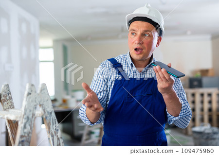 Worker in a protective helmet and blue overall discussing repair plan on the speakerphone of mobile phone Worker in a protective helmet and blue overall discussing repair plan on the speakerphone of mobile phone 109475960