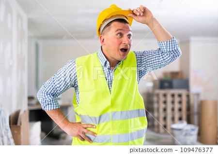 Frustrated worker in yellow vest and hard hat is dissatisfied with the way walls are plastered 109476377