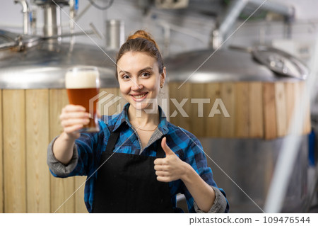 Portrait of female brewer who is standing with glass of beer on his workplace in brew-house Portrait of female brewer who is standing with glass of beer on his workplace in brew-house 109476544