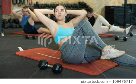 Motivated sporty young woman lying on her back and training core muscles during group training class in CrossFit center 109476717