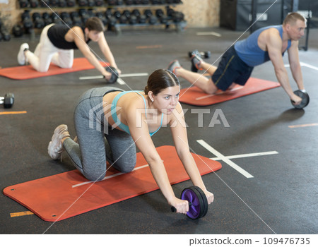 Motivated young woman practicing exercises with abs roller near other people in gym Motivated young woman practicing exercises with abs roller near other people in gym 109476735
