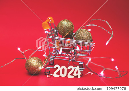 Christmas balls in shopping cart with garland and wooden numbers 2024 on red background. Christmas balls in shopping cart with garland and wooden numbers 2024 on red background. 109477976