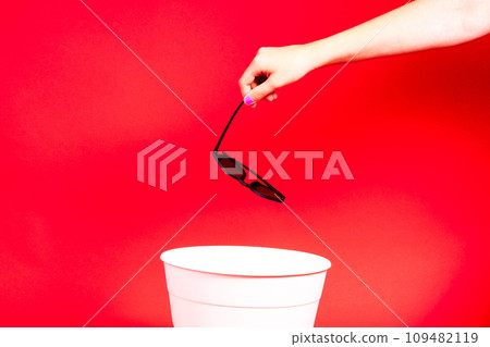 Hand throws sunglasses into trash bin with package on red background 109482119