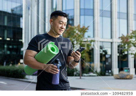 A young Asian sportsman man is standing on a city street in headphones, holding a phone, a yoga mat, and a bottle of water. A young Asian sportsman man is standing on a city street in headphones, holding a phone, a yoga mat, and a bottle of water. 109482959