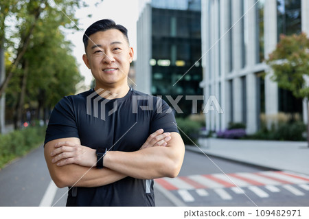 Portrait of a smiling young Asian male athlete standing on a city street, looking at the camera with his arms crossed. Portrait of a smiling young Asian male athlete standing on a city street, looking at the camera with his arms crossed. 109482971