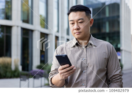 Close-up photo of a young Asian male businessman standing outside an office center with a phone in his hands, waiting for an appointment, making a call, typing a message. 109482979