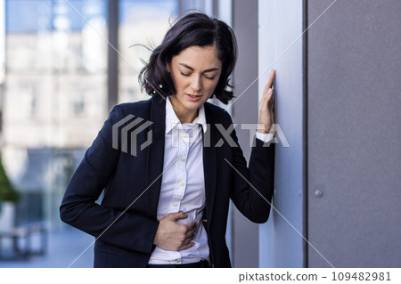 Young businesswoman suffering from stomach pain, standing near the office building, leaning against the wall, holding her body. 109482981