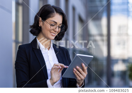 A smiling businesswoman is standing outside in an office center in a suit and using a tablet. Close-up photo. 109483025