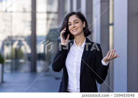 A young businesswoman is talking on the street near an office building on a mobile phone, discussing, gesturing with her hands. A young businesswoman is talking on the street near an office building on a mobile phone, discussing, gesturing with her hands. 109483046
