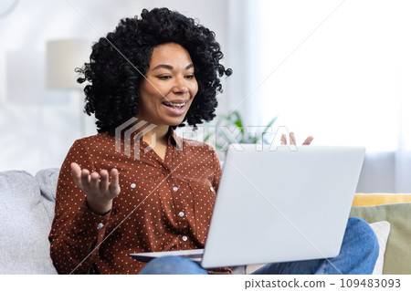 Close-up photo of a young African-American woman sitting on the couch at home and talking on a video call through a laptop, smiling, gesturing with her hands. 109483093