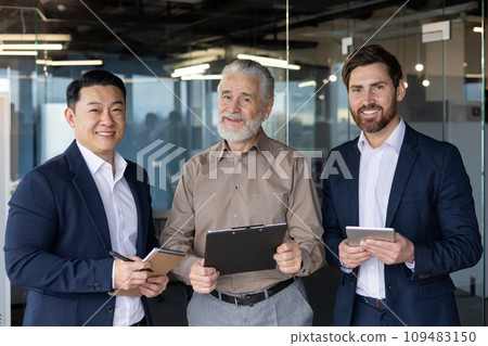 Portrait of three businessmen of different ages and nationalities. They are standing in the office, holding tablets and documents in their hands, confidently and smilingly looking at the camera. Portrait of three businessmen of different ages and nationalities. They are standing in the office, holding tablets and documents in their hands, confidently and smilingly looking at the camera. 109483150