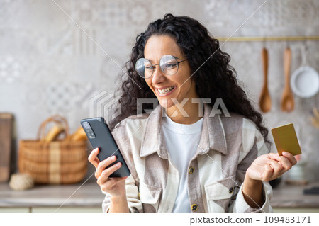 Young beautiful woman in kitchen with phone and bank credit debit card in hands, smiling Hispanic woman chooses gifts and products in online store, online shopping remotely from home. 109483171