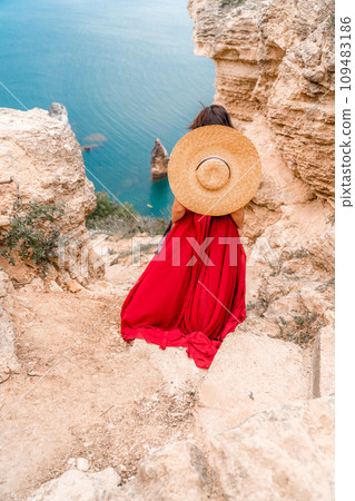 A girl with loose hair in a long red dress descends the stairs between the yellow rocks overlooking the sea. A rock can be seen in the sea. Sunny path on the sea from the rising sun A girl with loose hair in a long red dress descends the stairs between the yellow rocks overlooking the sea. A rock can be seen in the sea. Sunny path on the sea from the rising sun 109483186