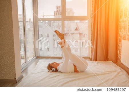 Side view portrait of relaxed woman listening to music with headphones lying on carpet at home. She is dressed in a white tracksuit. Side view portrait of relaxed woman listening to music with headphones lying on carpet at home. She is dressed in a white tracksuit. 109483297