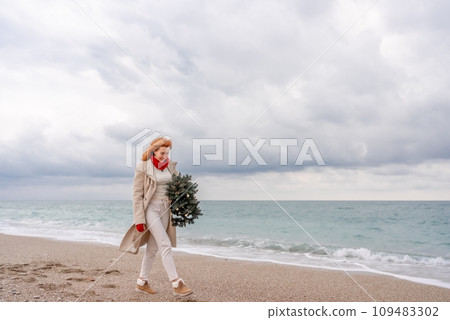 Redhead woman Christmas tree sea. Christmas portrait of a happy redhead woman walking along the beach and holding a Christmas tree in her hands. Dressed in a light coat, white suit and red mittens. Redhead woman Christmas tree sea. Christmas portrait of a happy redhead woman walking along the beach and holding a Christmas tree in her hands. Dressed in a light coat, white suit and red mittens. 109483302