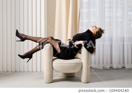 Playful young woman in a knitted dress with slit sits on an armchair 109484280