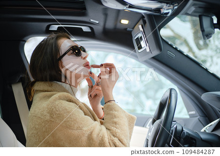 Woman applying lipstick wearing face mask while driving a car Woman applying lipstick wearing face mask while driving a car 109484287
