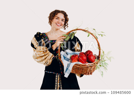 Portrait of smiling young adult woman dressed in a medieval dress holding a basket with vegetables brussels sprouts and apple fruits isolated white background Portrait of smiling young adult woman dressed in a medieval dress holding a basket with vegetables brussels sprouts and apple fruits isolated white background 109485600