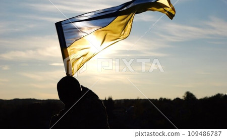 Silhouette of ukrainian army man stands at hill waving national banner against sunset. Male soldier lifting blue-yellow flag as symbol of victory against russian aggression. Fight for independence 109486787