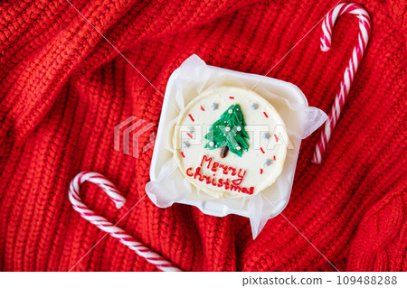 A festive Christmas cupcake with a white frosting and a green tree on top, on a red knitted background with candy canes. Merry Christmas lettering. 109488288