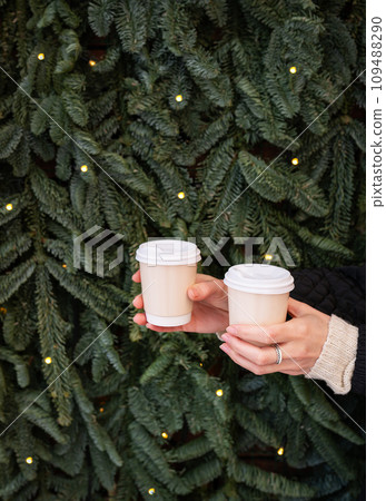 Two hands holding paper cups of coffee in front of a Christmas tree. 109488290