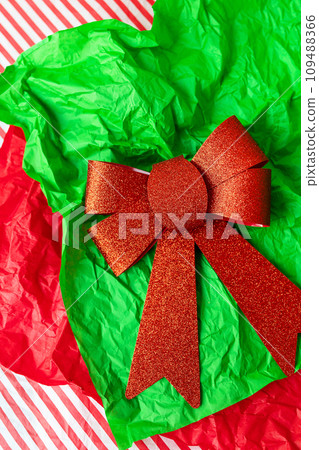 This is a photo of a red glitter bow on a green crumpled tissue paper with a red and white striped background. 109488366