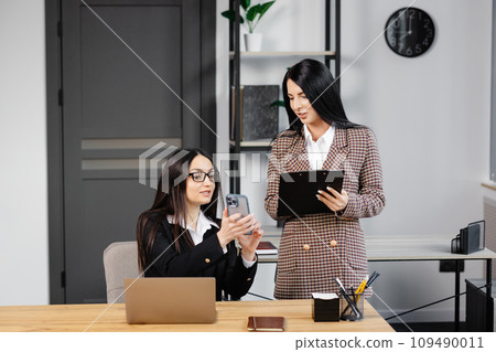 Two young attractive women are working in the office. A woman is talking on the phone, and her colleague is making notes in a notebook Two young attractive women are working in the office. A woman is talking on the phone, and her colleague is making notes in a notebook 109490011
