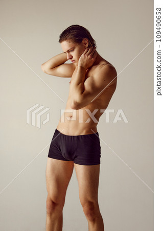 Handsome, shirtless, muscular young man fit fit body standing in underwear, holding hand on neck against grey studio background 109490658
