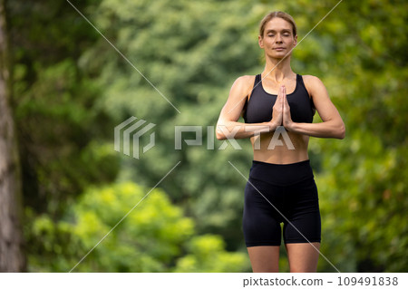 Adult slim woman in tight sportswear practicing yoga outdoors with prayer hands. Adult slim woman in tight sportswear practicing yoga outdoors with prayer hands. 109491838