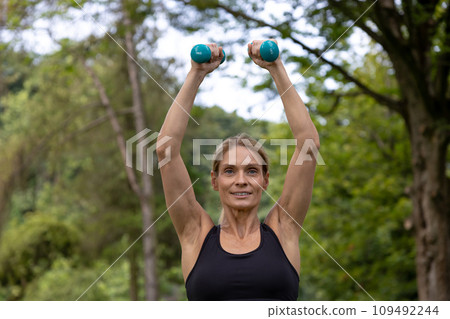 Sporty woman exercising with dumbbells outdoors has physical activity in park. Sporty woman exercising with dumbbells outdoors has physical activity in park. 109492244