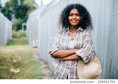 Confidently standing in a farm setting a female farmer in checkered attire smiles with crossed arms and a hat. Background of vibrant greenhouse tomatoes portrays care for nature and farm growth. Confidently standing in a farm setting a female farmer in checkered attire smiles with crossed arms and a hat. Background of vibrant greenhouse tomatoes portrays care for nature and farm growth. 109492768