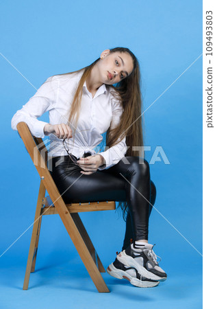 Pensive caucasian female with long fair hair wearing white shirt and leggings, holding glasses, tilting head to side, while looking at camera, isolated on blue studio background. Concept of posing. 109493803