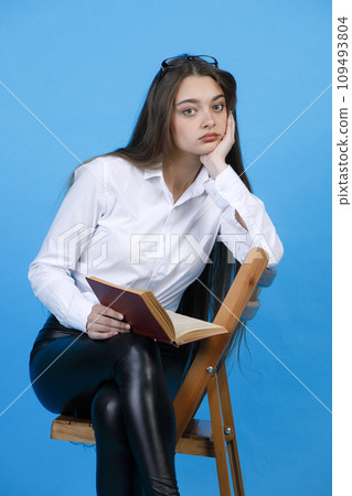 Bored caucasian woman with glasses on head, resting head on hand, while sitting on chair indoor. Portrait of upset girl with open book, while looking at camera, isolated on blue. Concept of emotions. 109493804
