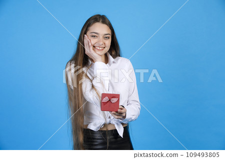 Portrait of cheerful young woman in blouse keeping hand on cheek from pleasure, while holding tiny gift box, decorated with bow and smiling at camera, isolated on blue background. Concept of emotions. Portrait of cheerful young woman in blouse keeping hand on cheek from pleasure, while holding tiny gift box, decorated with bow and smiling at camera, isolated on blue background. Concept of emotions. 109493805