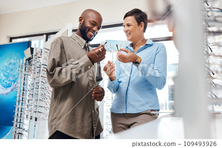 Eyesight, vision and optometry with a young man shopping for prescription glasses or eyewear with an optometrist or optician. Looking for spectacles with help from a female shop assistant from below 109493926