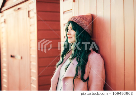 Stylish young smiling hipster woman with color hair wearing trendy peach color coat leaning on beach hut and enjoying moment. Urban seasonal city street fashion. Color of 2024 year. Selective focus. 109493968