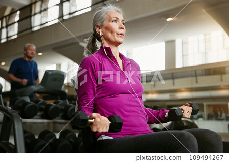 Fitness, dumbbell and senior woman at a gym for training, wellness and cardio with earphones, music or mindset. Weightlifting, bodybuilding and elderly female person at sports center for arm workout Fitness, dumbbell and senior woman at a gym for training, wellness and cardio with earphones, music or mindset. Weightlifting, bodybuilding and elderly female person at sports center for arm workout 109494267