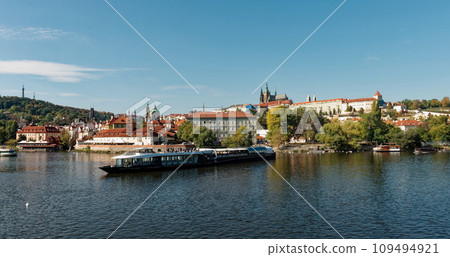 A boat is sailing on a river with a cityscape in the background. Pleasure boat on the Vltava pier in the fall. View of Prague. A boat is sailing on a river with a cityscape in the background. Pleasure boat on the Vltava pier in the fall. View of Prague. 109494921