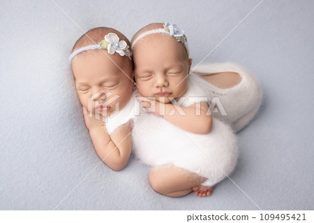 Tiny newborn twin girls. A newborn twin embraces and sleeps on his sister, Newborn twin girls against a pale blue blanket with white bandages and white delicate woolen bodysuits.  109495421