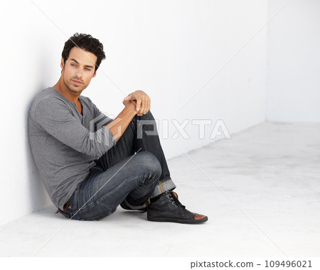 Handsome, fashion and young man by a white wall with casual, cool and trendy outfit. Attractive, confidence and serious male model from Canada sitting on floor with edgy style for aesthetic. 109496021