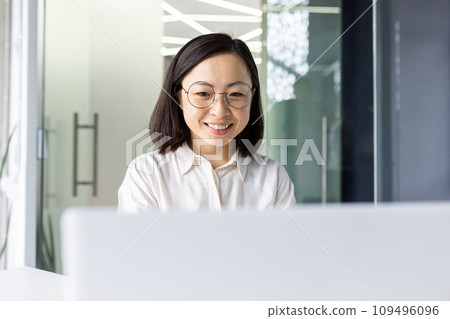 Successful Asian business woman at workplace inside office, female employee smiling contentedly using laptop at work, woman in light clothes satisfied with achievement results, typing on keyboard. Successful Asian business woman at workplace inside office, female employee smiling contentedly using laptop at work, woman in light clothes satisfied with achievement results, typing on keyboard. 109496096