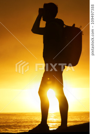 Silhouette, man and hiking at ocean with sunset, backpack and scenery for holiday or vacation is summer. Shadow, person and relaxing with view, adventure and hiker for recreation, travel or sun flare 109497388