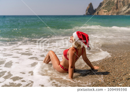 Female beach Santa hat wave coast. beach relaxation seaside. A woman in a red swimsuit enjoying her time on the beach, sits on the sand and being covered by a wave. Female beach Santa hat wave coast. beach relaxation seaside. A woman in a red swimsuit enjoying her time on the beach, sits on the sand and being covered by a wave. 109499398