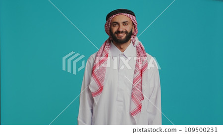 Middle eastern adult in religious attire smiling and posing proudly, representing muslim cultural identity with red and white print headscarf and robe. Young confident islamic person. Middle eastern adult in religious attire smiling and posing proudly, representing muslim cultural identity with red and white print headscarf and robe. Young confident islamic person. 109500231