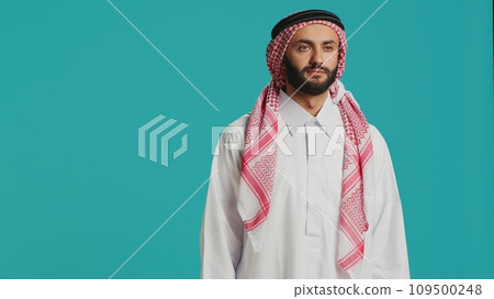 Middle eastern man in traditional attire smiling and posing with confidence, representing arabic cultural identity with checkered print headscarf and white robe. Young cheerful arab person. 109500248
