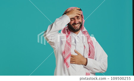 Cheerful person laughing at a joke in studio, having fun while he wears muslim traditional clothing and kufiyah. Young arab man enjoying laugh with people, expressing happiness and joy. Cheerful person laughing at a joke in studio, having fun while he wears muslim traditional clothing and kufiyah. Young arab man enjoying laugh with people, expressing happiness and joy. 109500255