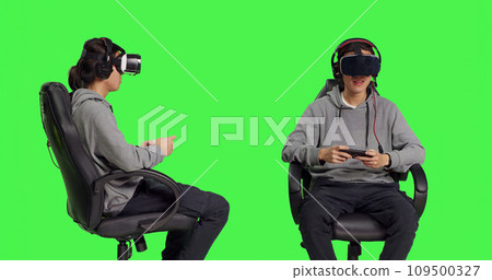 Person plays videogames with vr glasses and phone, participating in online multiplayer competition sitting on chair over greenscreen backdrop. Asian man having fun with action gameplay. Person plays videogames with vr glasses and phone, participating in online multiplayer competition sitting on chair over greenscreen backdrop. Asian man having fun with action gameplay. 109500327
