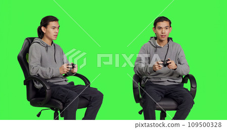 Asian guy playing videogames over greenscreen backdrop, using controller and having fun with friends on online gaming competition. Young man gamer enjoying rpg gameplay, blank background. Asian guy playing videogames over greenscreen backdrop, using controller and having fun with friends on online gaming competition. Young man gamer enjoying rpg gameplay, blank background. 109500328