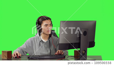 Asian gamer on desktop at workstation, playing internet videogames with others. Young man having fun with role playing battle, communicating with friends over greenscreen template. 109500338